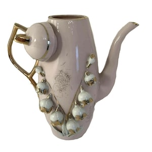 May include: A pink ceramic teapot with a gold handle, spout, and lid. The teapot is decorated with white and gold lily of the valley flowers. The teapot is a vintage style.