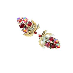 May include: A pair of vintage leaf-shaped earrings. The earrings feature a gold-tone metal design with red and iridescent crystal accents. The crystals are clustered to resemble berries. The earrings are set against a white background.