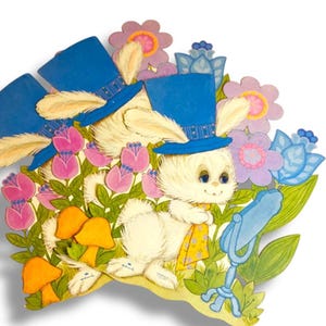 May include: A vintage Easter-themed die-cut decoration featuring a white bunny wearing a blue hat and yellow scarf, surrounded by pink and purple flowers, orange mushrooms, and blue accents. The bunny is smiling.