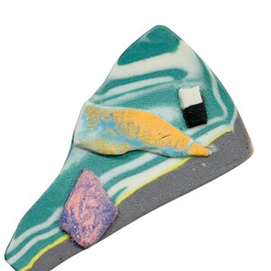 May include: A colorful, abstract brooch with a unique design. The brooch features a teal, white, and gray base with yellow, orange, and pink accents. A small black and white square is also present. The brooch is a unique piece of wearable art.