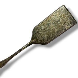 May include: Silver-toned serving utensil with an ornate, rectangular blade and a long, slender handle. The blade features an engraved floral pattern. The handle has a decorative end with a shell-like design. The utensil is likely vintage.