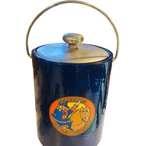 May include: A navy blue ice bucket with a gold-colored handle and lid. The bucket features a circular logo with the text "Schweppes" and the phrase "Curiously Refreshing."
