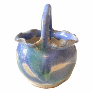 May include: A handmade ceramic basket with a handle. The basket features a blue, green, and beige color scheme. The rim of the basket has a wavy design. The handle is arched and attached to the sides of the basket.