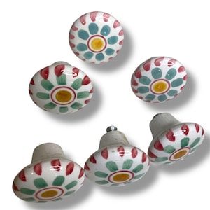 May include: A collection of six ceramic cabinet knobs. Each knob features a white base with a floral design in red, green, and blue, centered with a yellow circle. The knobs are in various orientations, showcasing their decorative patterns.