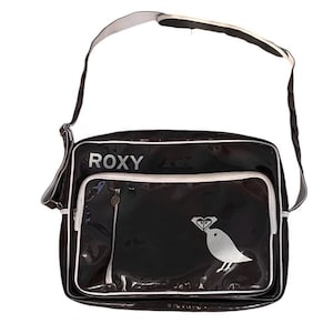 May include: Black Roxy shoulder bag with a white trim and strap. The bag features a silver zipper and a silver bird graphic with a heart-shaped diamond. The word "ROXY" is printed in white on the top of the bag.