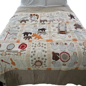 May include: A handmade quilt with a beige and cream background, featuring various animal and object appliqués in brown, orange, and red. The quilt is on a bed with white pillows. The quilt's design includes squares and borders.