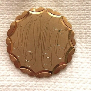 May include: A gold-coloured, circular brooch with a scalloped edge. The brooch features an engraved monogram of three stylized letters. The metal has a warm, polished finish. The design is classic and elegant.