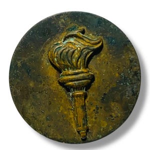 May include: A round, antique-style bronze emblem featuring a torch with a flame. The torch is centered on the emblem, with a detailed design. The emblem has a weathered appearance, with a dark bronze color and a slightly raised relief.