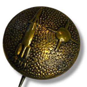 May include: A round, antique-style pin with a textured surface and a gold finish. The pin features a rocket ship and a satellite design. The pin has a long, sharp needle on the back for attachment.