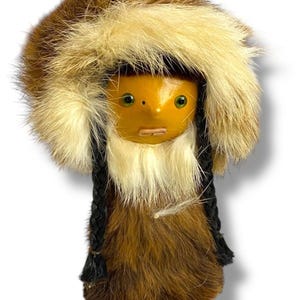 Puede incluir: Muñeca artesanal con rostro de madera y ropa de piel. La muñeca tiene una capucha forrada de piel, ojos verdes y trenzas negras. La ropa de la muñeca es de piel marrón y beige.