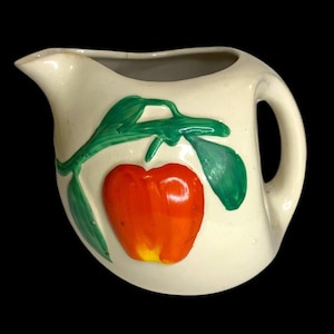 May include: Cream ceramic jug with a handle and spout. It features a raised, bright red apple and green leaves design. A delightful piece for serving drinks or as a decorative item.
