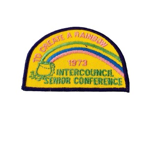 K&ouml;nnte beinhalten: Ein gestickter Aufn&auml;her in Gelb, Blau, Gr&uuml;n und Rosa mit einem Regenbogen und dem Text "To Create A Rainbow 1973 Intercouncil Senior Conference".