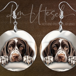 May include: A pair of round earrings featuring a brown and white dog illustration. The dog's head and paws are visible, with a white background and silver-colored ear wires. The earrings have a detailed, artistic design.