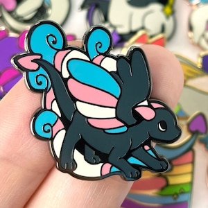 Puede incluir: Un pin de esmalte negro y blanco con un dragón con alas translúcidas azules, rosas y blancas. El dragón tiene una cola enroscada y una cinta rosa alrededor de la cabeza.