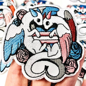 May include: White embroidered patch with two white dragons with blue and pink wings holding a transgender pride flag. The dragons are surrounded by pink and blue flowers.