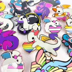 May include: A collection of enamel pins featuring a cartoon dragon design with various pride flag color schemes. The pins have gold-colored metal borders and include designs with rainbow hearts, purple wings, and other pride symbols.