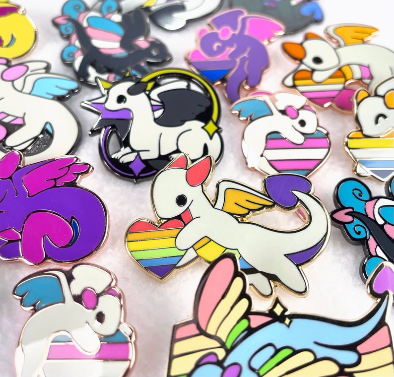 May include: A collection of enamel pins featuring a cartoon dragon design with various pride flag color schemes. The pins have gold-colored metal borders and include designs with rainbow hearts, purple wings, and other pride symbols.