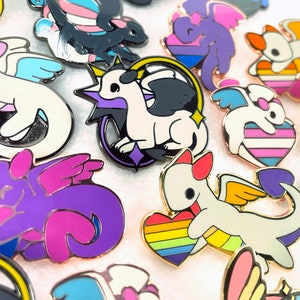 May include: A collection of enamel pins featuring stylized dragons with various pride flag color schemes. The pins have gold-colored borders and depict dragons with wings, hearts, and other decorative elements. The dragons are in white, black, purple, and blue.