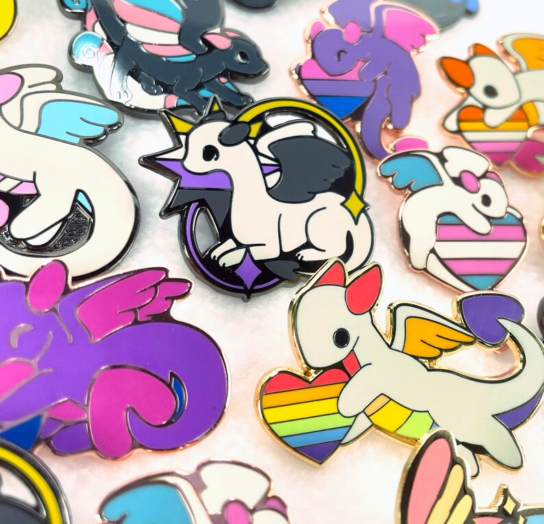 May include: A collection of enamel pins featuring stylized dragons with various pride flag color schemes. The pins have gold-colored borders and depict dragons with wings, hearts, and other decorative elements. The dragons are in white, black, purple, and blue.