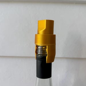 May include: A gold-coloured cylindrical bottle stopper with a stepped design, featuring a textured finish. The stopper is fitted over the threaded neck of a glass bottle, which has a black sleeve covering the lower portion of the bottle neck.