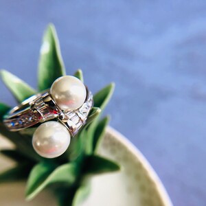 May include: A silver ring with two large, round, white pearls. The ring is adorned with smaller, clear gemstones. The ring is displayed on a green plant with a blue background.