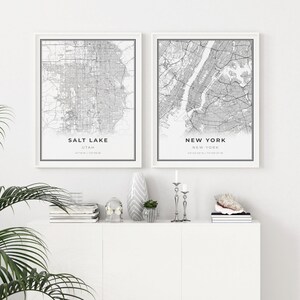Puede incluir: Dos mapas en blanco y negro enmarcados de Salt Lake City, Utah y Nueva York, Nueva York. Los mapas est&aacute;n colgados en una pared blanca sobre un estante blanco con art&iacute;culos decorativos.