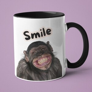 Op de afbeelding: Witte keramische koffiemok met een zwarte rand en handvat. De mok heeft een glimlachende chimpansee met het woord "Smile" in zwart boven de afbeelding.