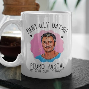 Könnte beinhalten: Weiße Keramik-Tasse mit Henkel, mit einer Cartoon-Illustration von Pedro Pascal in einem rosa herzförmigen Design. Auf der Tasse steht der Text "MENTALLY DATING PEDRO PASCAL MY COOL SLUTTY DADDY". Im Hintergrund ist eine Kaffeemaschine zu sehen.