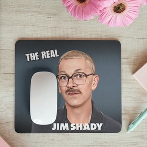 Peut inclure: Un tapis de souris noir avec une bordure blanche. Le tapis de souris présente une photo d'un homme avec une moustache et des lunettes. Le texte "THE REAL JIM SHADY" est imprimé sur le tapis de souris.
