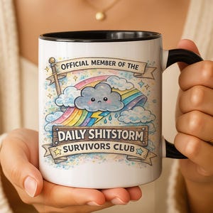 May include: White ceramic mug with a black interior and handle. The mug features a colourful illustration of a rainbow, cloud, and lightning bolt, with the text "OFFICIAL MEMBER OF THE DAILY SHITSTORM SURVIVORS CLUB."