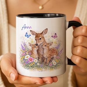 May include: White ceramic mug with a black interior and handle. The mug features a watercolour illustration of a deer family with two fawns amidst colourful flowers and butterflies. The name "Anna" is written in script.