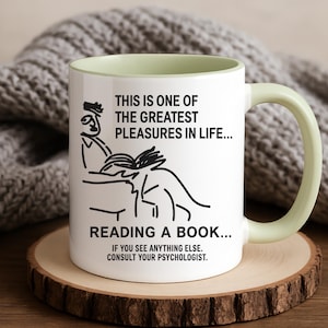 Pode incluir: Caneca de cerâmica branca com alça e interior verde claro. A caneca apresenta um desenho a traço preto de uma pessoa lendo um livro, com o texto "THIS IS ONE OF THE GREATEST PLEASURES IN LIFE... READING A BOOK..."