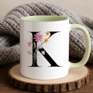 Puede incluir: Taza de cerámica blanca con la letra "K" negra adornada con flores rosas, naranjas y moradas. La taza tiene un interior y un asa de color verde claro, colocada sobre un posavasos de madera. Una manta de punto gris está en el fondo.