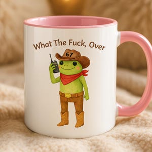 May include: White ceramic mug with a pink interior and handle. The mug features a cartoon frog dressed as a cowboy, holding a walkie-talkie, with the text "What The Fuck, Over" and the number 67.
