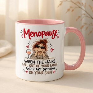 May include: A white ceramic mug with a light pink interior and handle. The front features a cartoon illustration of a person with messy hair and sunglasses, a wine glass, and the text: Menopause WHEN THE HAIRS FALL OUT OF YOUR FANNY AND START GROWING ON YOUR CHIN.