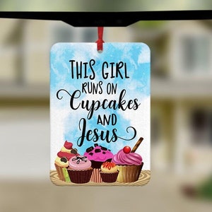 Könnte beinhalten: Ein weißer Auto-Lufterfrischer mit blauem Aquarellhintergrund. Der Text lautet "This girl runs on cupcakes and Jesus". Das Bild zeigt eine Vielzahl von bunten Cupcakes.