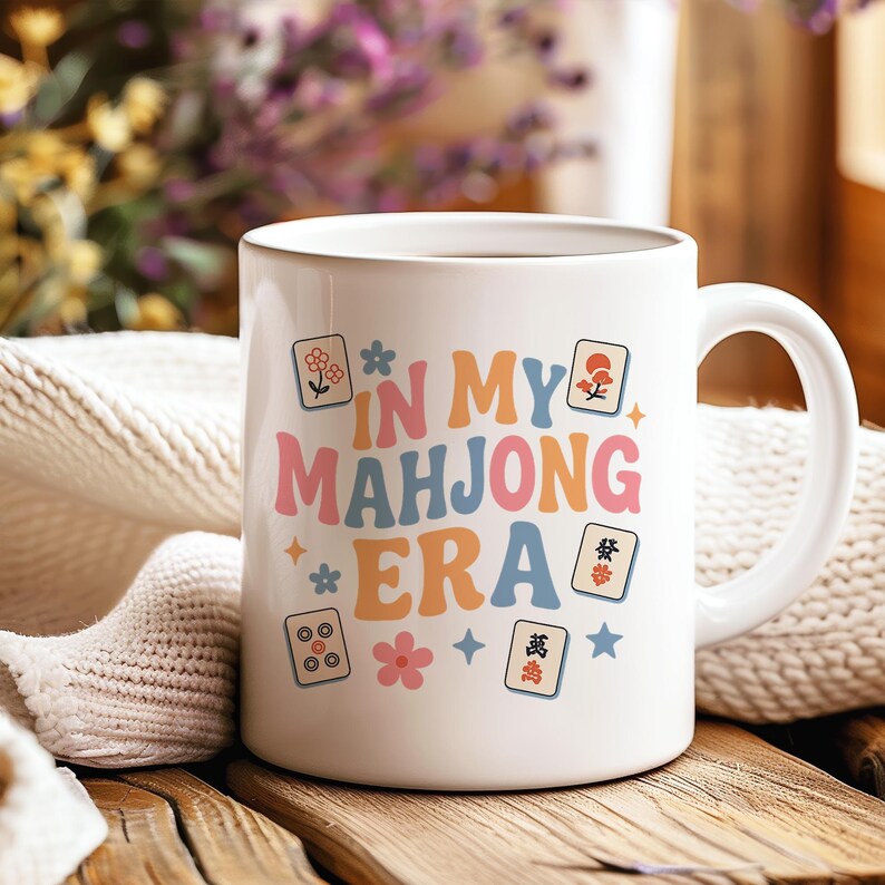 May include: White ceramic mug with the text "IN MY MAHJONG ERA" in colorful retro font. The mug features various Mahjong tile illustrations and floral accents, perfect for a game enthusiast.