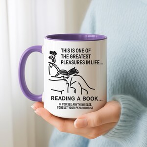May include: White ceramic mug with a purple interior and handle. The mug features a black line drawing of a person reading a book, with text that reads, "THIS IS ONE OF THE GREATEST PLEASURES IN LIFE... READING A BOOK... IF YOU SEE ANYTHING ELSE, CONSULT YOUR PSYCHOLOGIST."