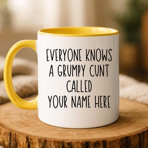 Può includere: Tazza in ceramica bianca con manico e interno gialli. La tazza presenta il testo "EVERYONE KNOWS A GRUMPY CUNT CALLED YOUR NAME HERE" in nero, in grassetto. La tazza è appoggiata su una superficie di legno.