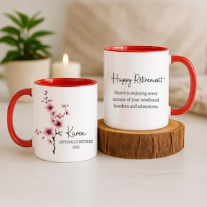 May include: Two white ceramic mugs with red interiors and handles. One mug features a floral design and the text "Karen OFFICIALLY RETIRED 2025." The other mug says "Happy Retirement" with a message below. Both mugs are on a wooden surface.
