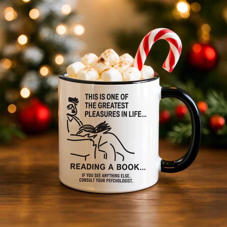 May include: White ceramic mug with a black handle and rim, filled with marshmallows and a candy cane. The mug has a graphic of a person reading and the text: "THIS IS ONE OF THE GREATEST PLEASURES IN LIFE... READING A BOOK... IF YOU SEE ANYTHING ELSE. CONSULT YOUR PSYCHOLOGIST."