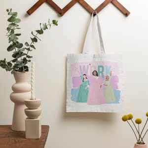 Op de afbeelding: Een witte canvas tote bag met een roze, blauwe en groene illustratie van drie vrouwen in jurken. De tekst "WORK" is in groen gedrukt boven de vrouwen. De tas hangt aan een houten wandrek.