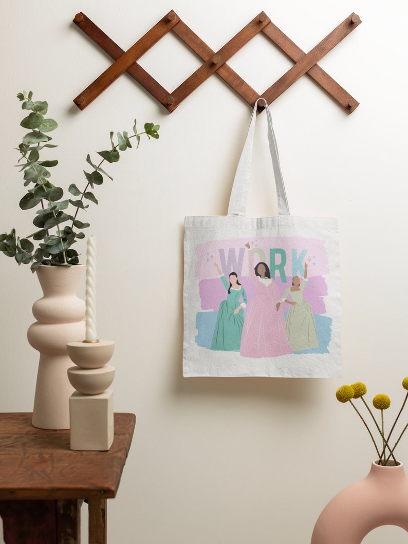 Op de afbeelding: Een witte canvas tote bag met een roze, blauwe en groene illustratie van drie vrouwen in jurken. De tekst "WORK" is in groen gedrukt boven de vrouwen. De tas hangt aan een houten wandrek.