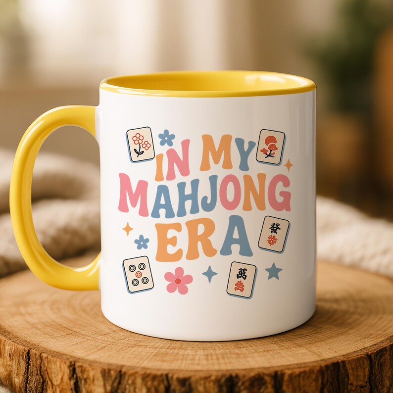 May include: White ceramic mug with a yellow handle and interior. The mug features the text "IN MY MAHJONG ERA" in colorful letters, along with floral and Mahjong tile graphics. The mug is set on a wooden surface.