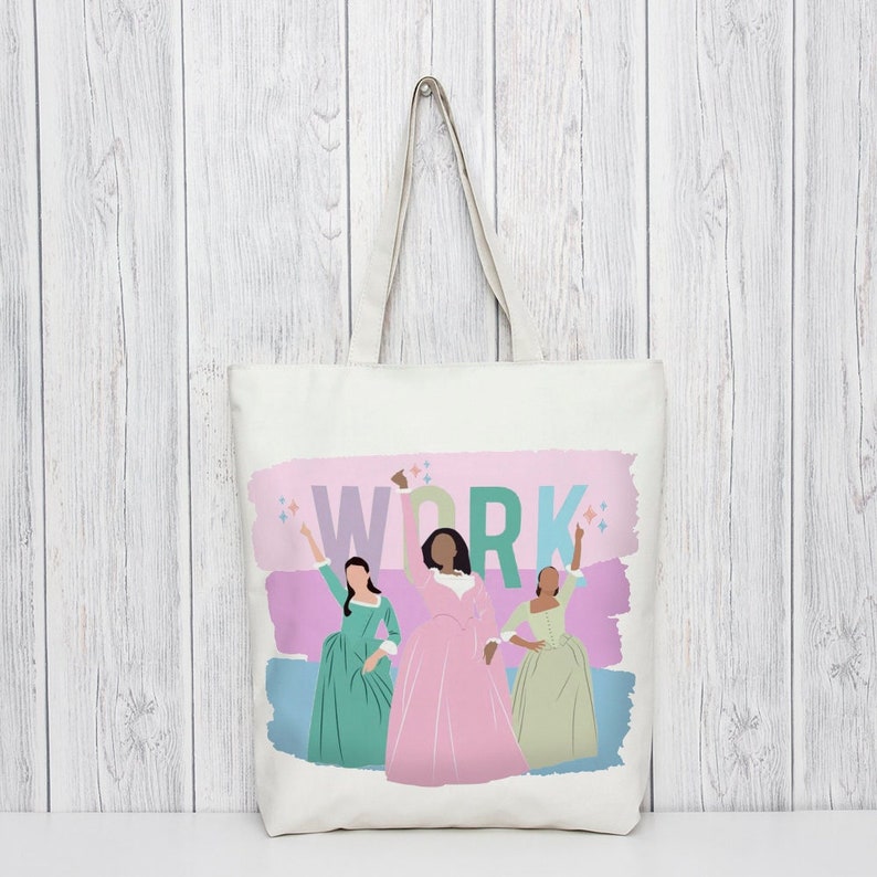 Op de afbeelding: Een witte canvas tote bag met een roze en blauwe aquarel achtergrond. De woorden "WORK" zijn gedrukt in grote, kleurrijke letters. Drie vrouwen in kleurrijke jurken zijn afgebeeld op de achtergrond.
