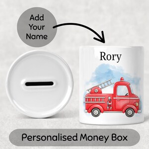 May include: White ceramic money box with a slot for coins, next to a cylindrical bank decorated with a red fire truck design and the name "Rory". The text "Add Your Name" and "Personalised Money Box" are also visible.