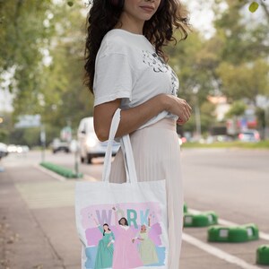 Op de afbeelding: Een witte tote bag met een roze, groene en blauwe illustratie van drie vrouwen in jurken en het woord "WORK" in roze letters. De tas wordt gedragen door iemand die een wit T-shirt, een roze rok en een bril draagt.