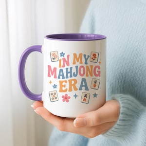 May include: White ceramic mug with a purple handle and interior. The mug features the text "IN MY MAHONG ERA" in colorful retro font, along with Mahjong tiles and floral graphics. A fun gift for Mahjong players.