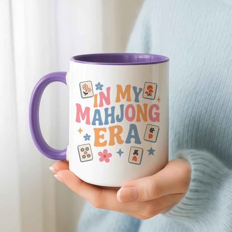 May include: White ceramic mug with a purple handle and interior. The mug features the text "IN MY MAHONG ERA" in colorful retro font, along with Mahjong tiles and floral graphics. A fun gift for Mahjong players.