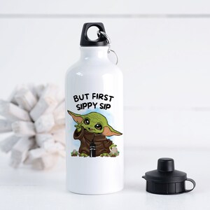 May include: White aluminum water bottle with a black lid and a black carrying loop. The bottle has a graphic of a green alien character holding a smaller bottle with the text "But First Sippy Sip".